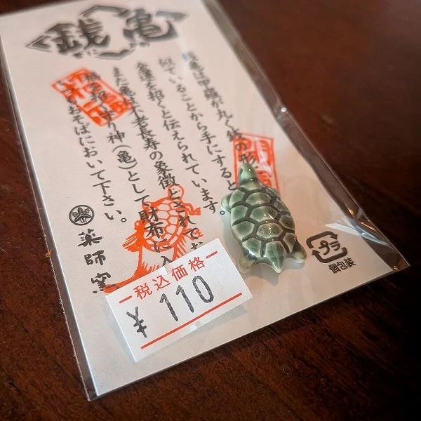 わぁ〜かわいらしいですねぇ🐢✨
銭亀（ぜにがめ）」の陶製お守り。亀は昔から長寿や金運の象徴とされていて、「銭亀」は特に“お金を呼ぶ亀”として縁起がいいんですよ。
小さなサイズで、陶器ならではの艶やかでしっとりした質感も上品。緑がかった釉薬の色合いも美しくて、つるんとした背中に小さな六角模様がちゃんと入っているのも職人さんの丁寧な仕事ぶりが伝わります。
税込110円というお手頃価格もうれしいですね。お財布に忍ばせたり、玄関やデスクにちょこんと置いたりするのもおすすめです。「金運アップ」「長寿祈願」「厄除け」として、お守り感覚で持つ方が多いですよ。
あなたなら、この子をどこに飾りたいですか？